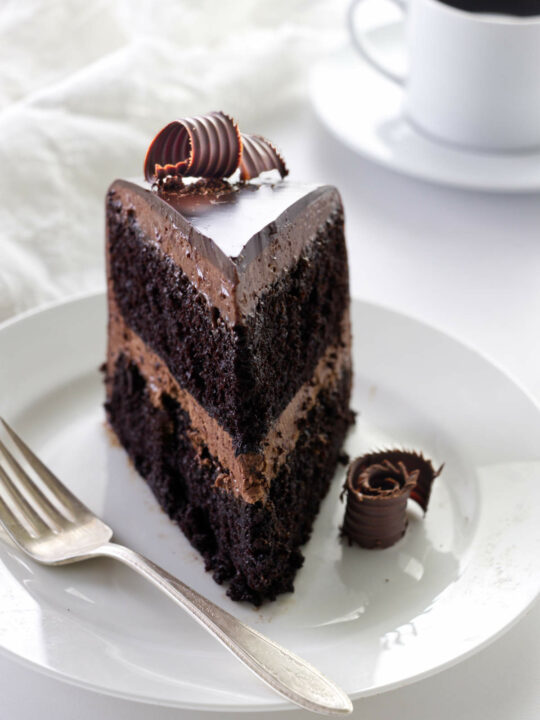 A slice of chocolate ganache cake decorated with chocolate curls on a plate with a fork, a napkin and cup of coffee in the background.
