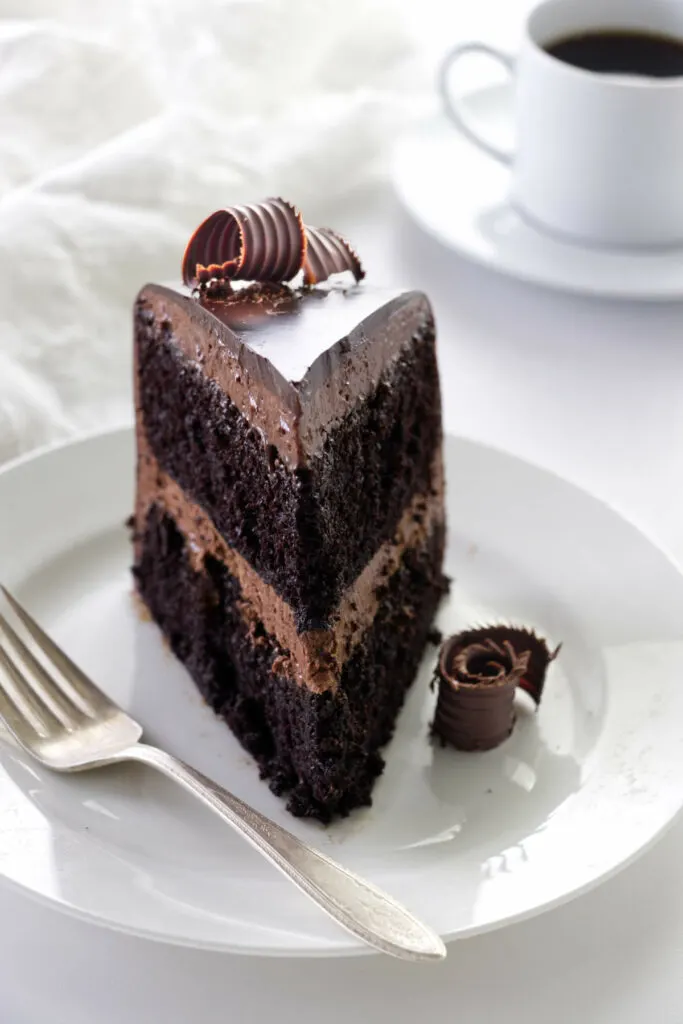 A slice of chocolate ganache cake decorated with chocolate curls on a plate with a fork, a napkin and cup of coffee in the background.