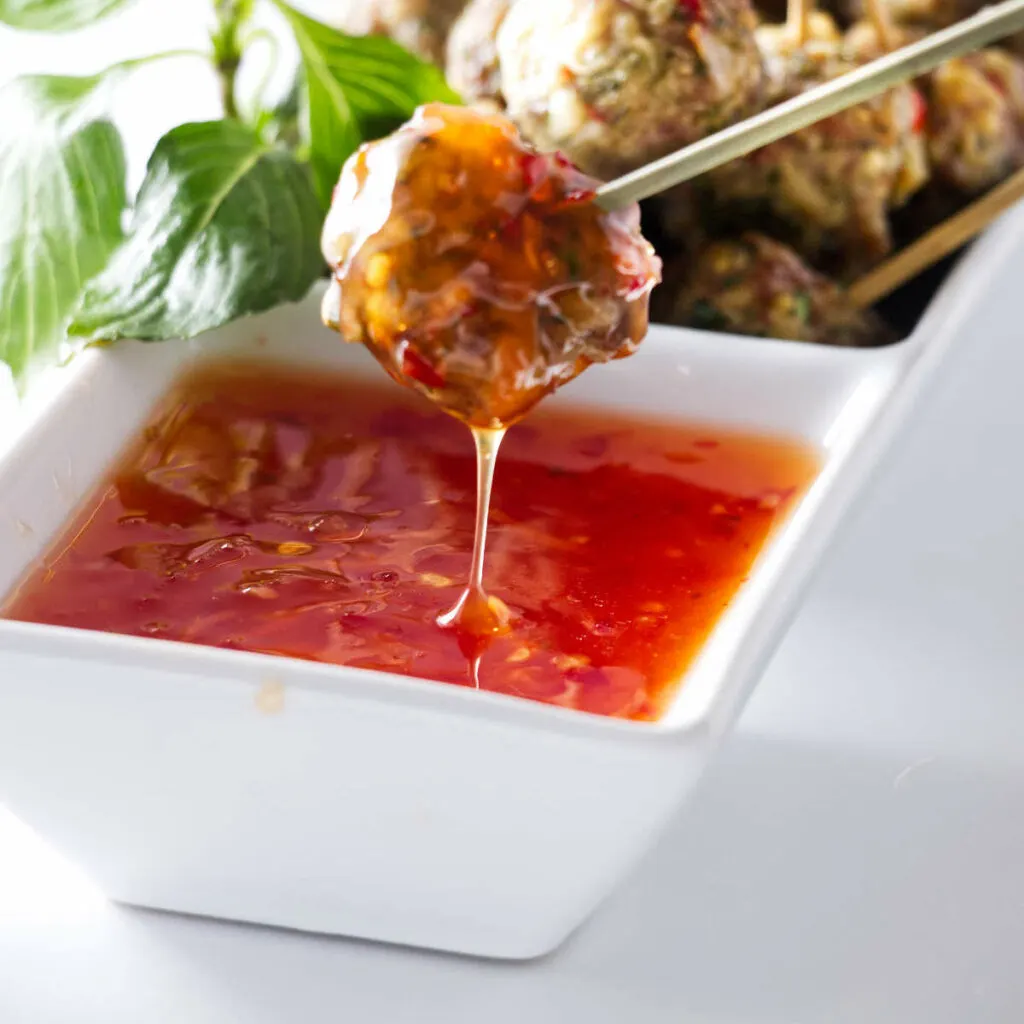 A meatball appetizer on a pick being dipped in a sweet/savory chili sauce.