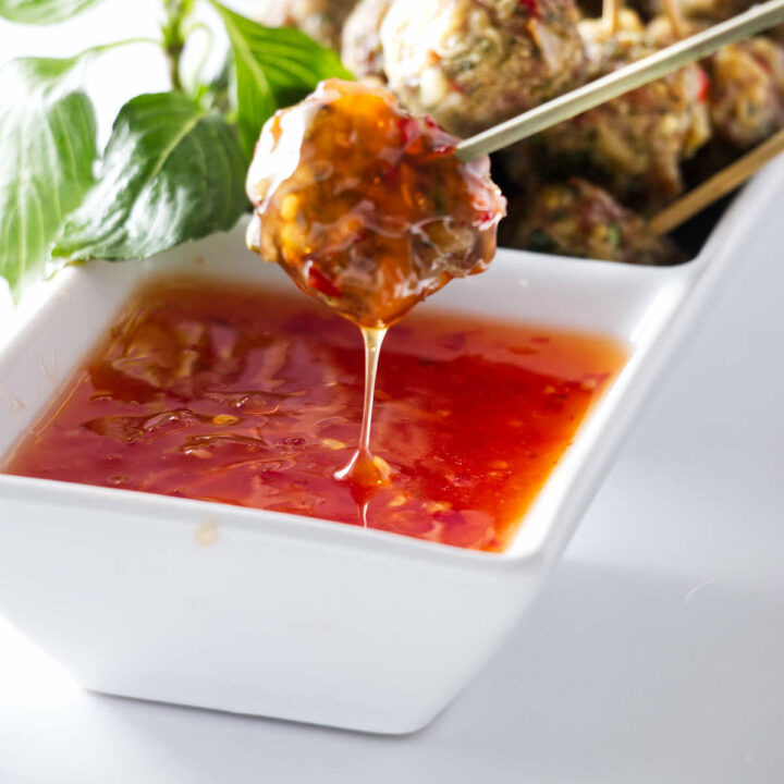 A meatball appetizer on a pick being dipped in a sweet/savory chili sauce.