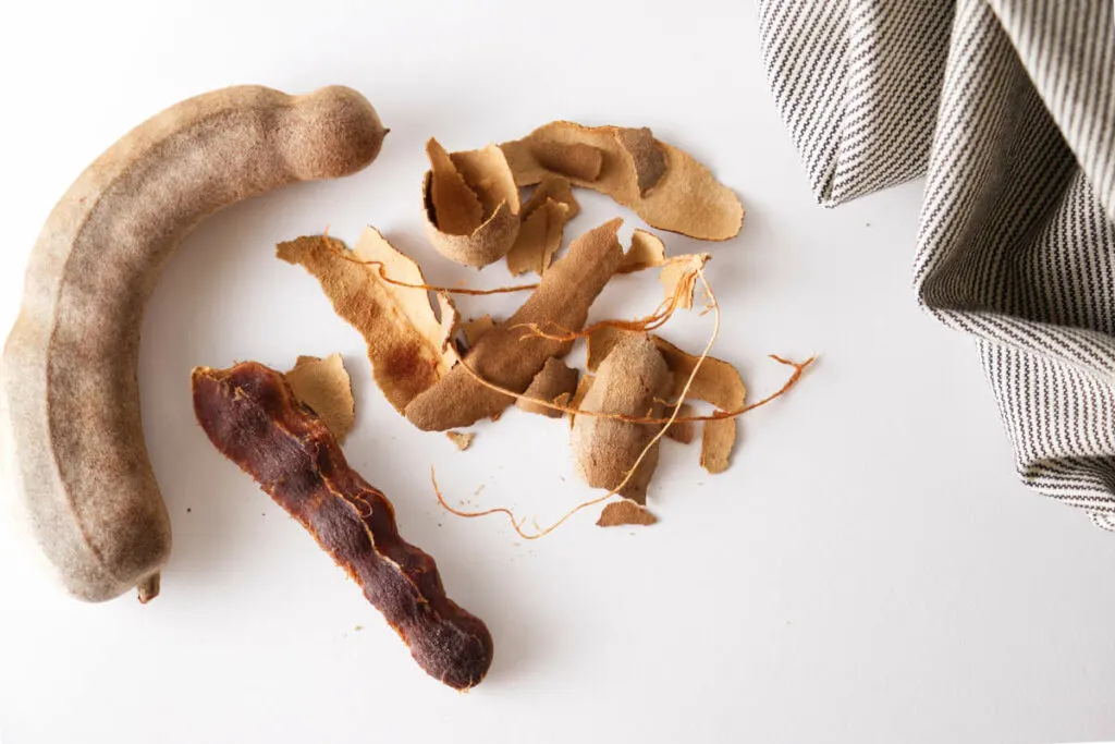 Two dried tamarind pods,One whole unhusked tamarine and one with the husk and strings removed exposing the inner portion.
