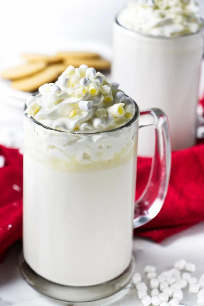 Two mugs of white hot chocolate with cookies in the background.