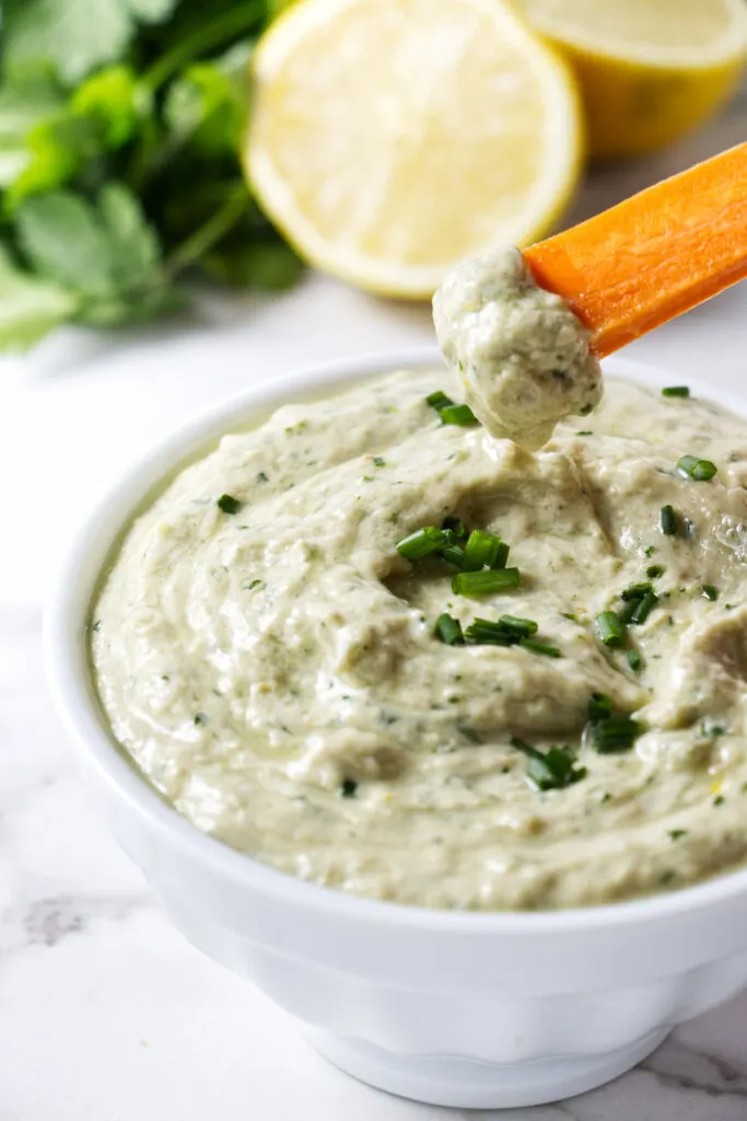 Dipping a carrot stick in lemon herb tahini dip with herbs.
