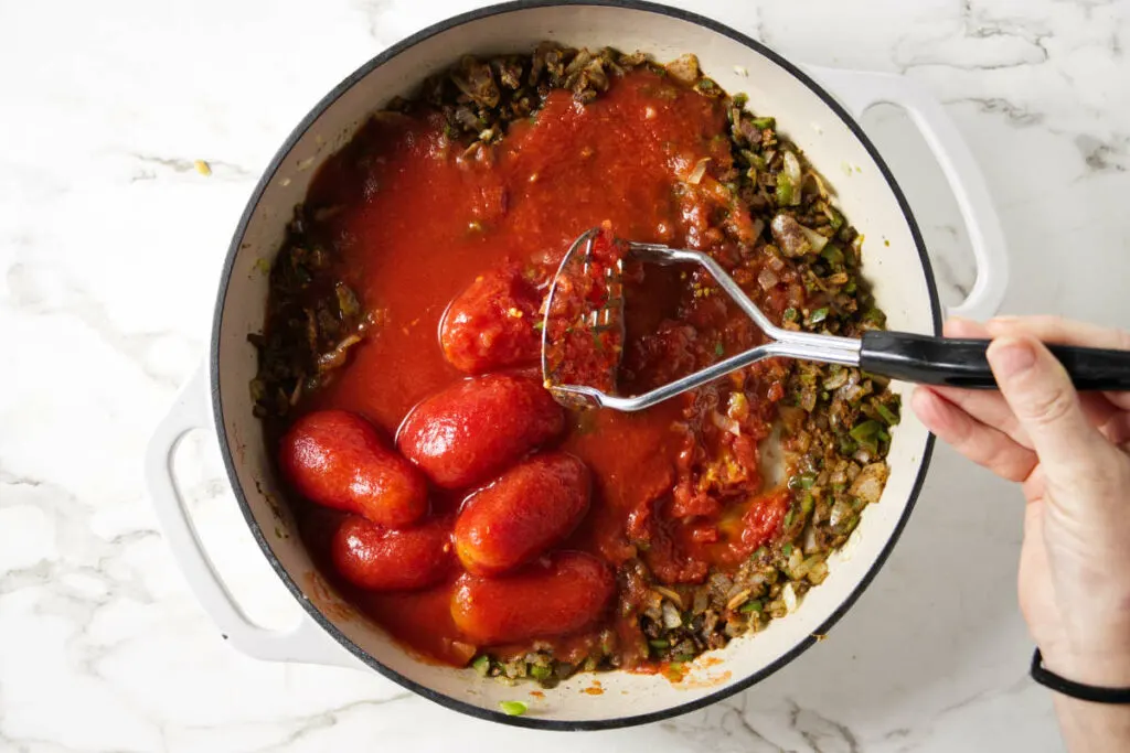 Adding the canned tomatoes and crushing them with a potato masher.