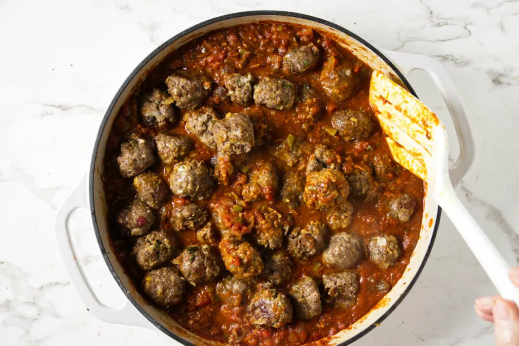 Stirring lamb meatballs into a skillet with tomato sauce.