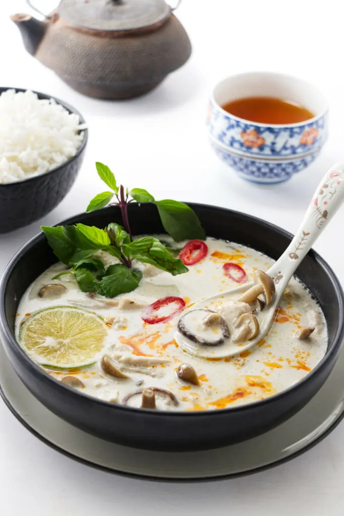 Tom Kha Soup in a dish with a dish of rice, tea and teapot in the background.