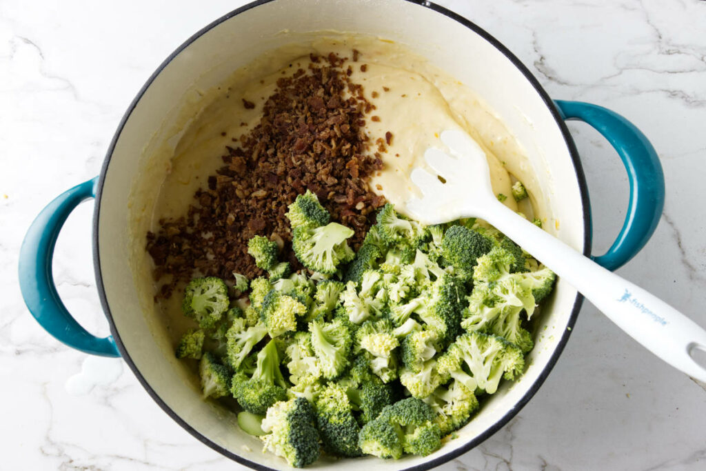 Adding broccoli and bacon to cheese sauce in a pot.