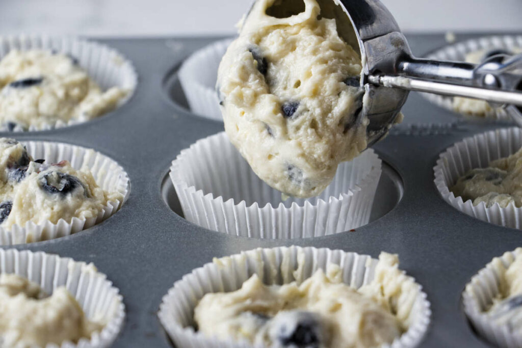 Scooping blueberry muffin batter into a muffin tin.