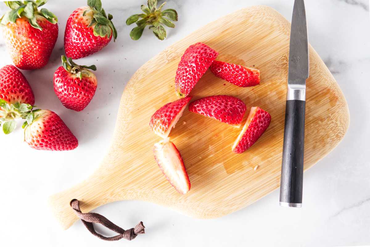 A cutting board, knife and sliced strawberries, fresh strawberries are next to the cutting board.