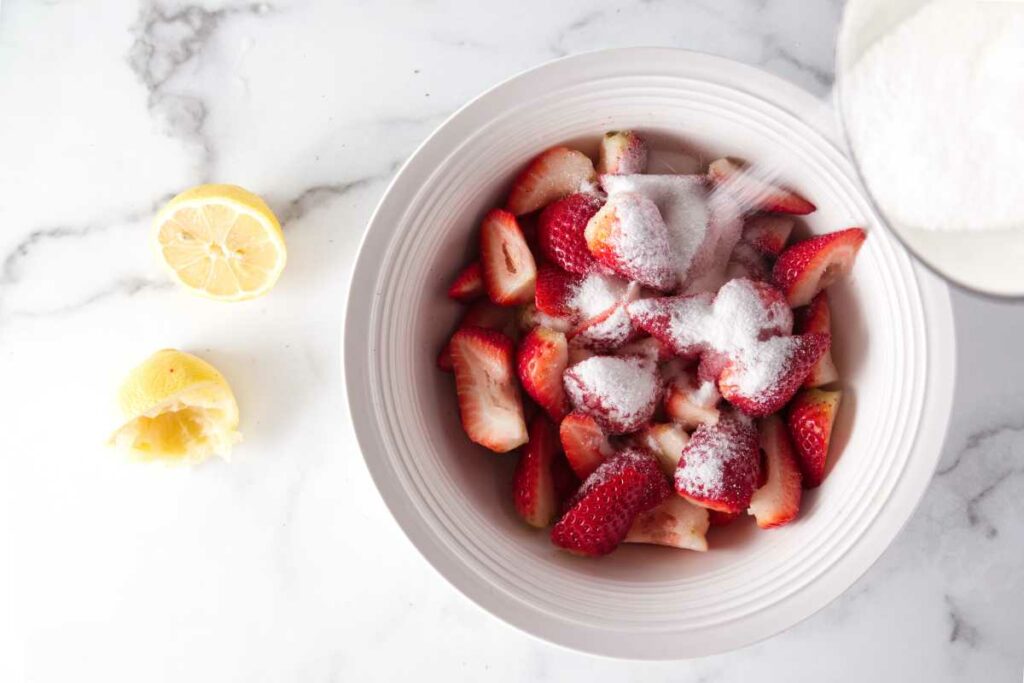 A bowl of freshly sliced strawberries with sugar being poured over them. Lemon halves next to the bowl.