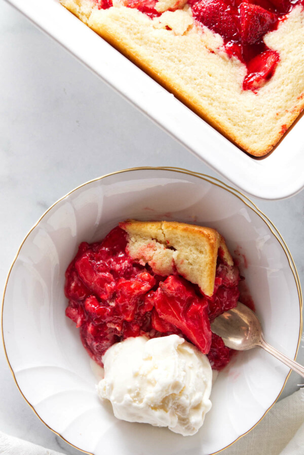 A serving of strawberry spoon cake with a scoop of vanilla ice cream. The dish of spoon cake next to the serving.