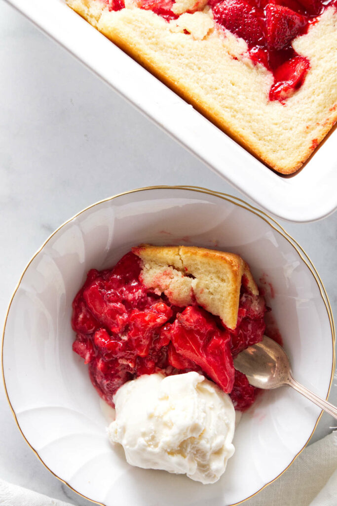 A serving of strawberry spoon cake with a scoop of vanilla ice cream. The dish of spoon cake next to the serving.
