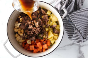 Adding root veggies, oxtail meat, and broth to a pot of soup.