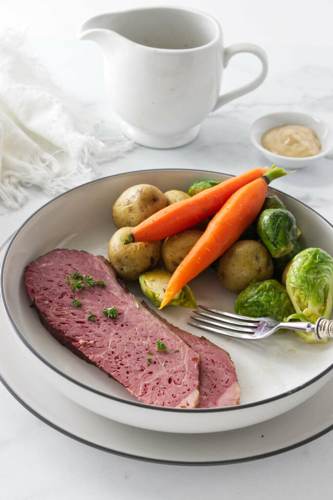 A serving of sous vide corned beef with vegetables. A pitcher of hot broth in the background.