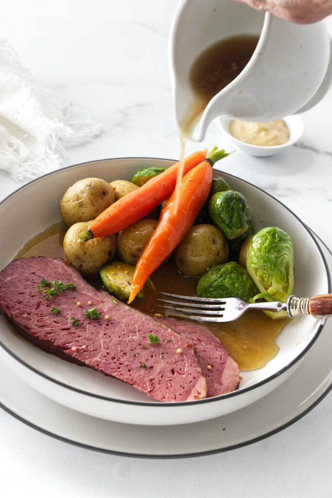 A serving of sous vide corned beef, carrots, potatoes and brussels sprouts. Pitcher of hot broth being poured over the top.
