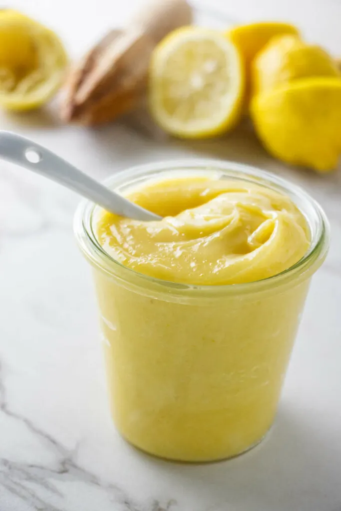 Thick lemon curd in a jar with several lemons in the background.