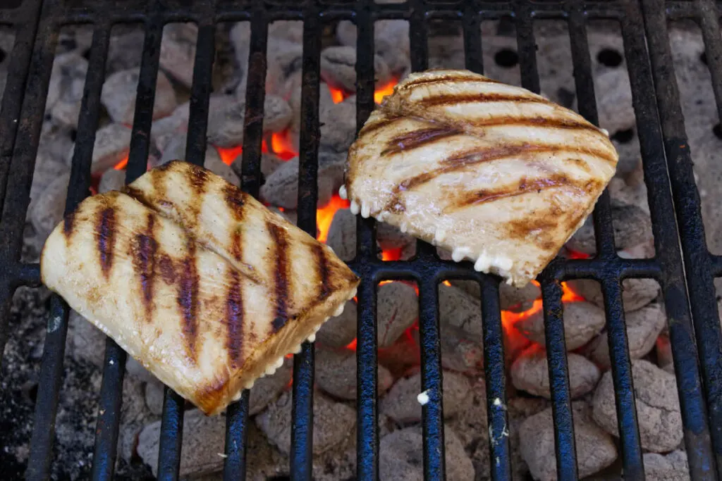 Two Mahi Mahi fillets on a grill grate over hot coals.