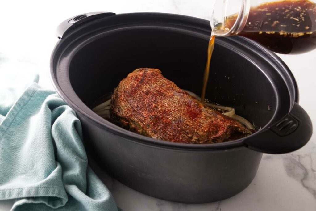 Pouring broth into a slow cooker with a tri tip and onions.