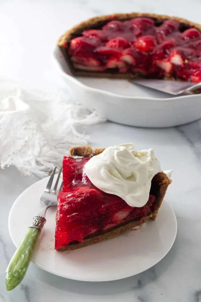 A slice of strawberry pie topped with whipped cream in front of a pie dish.