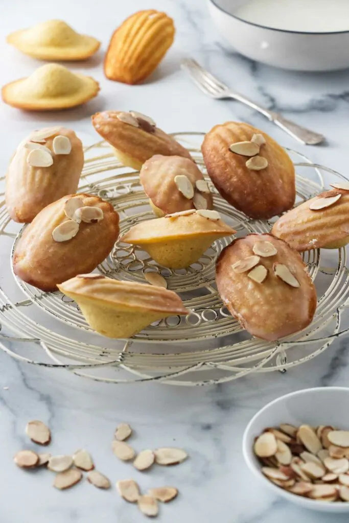 glazed almond madeleines drying on a wire rack. A dish of sliced almonds in the foreground, 3 unglazed madeleines, a fork and a dish of glaze in the background.