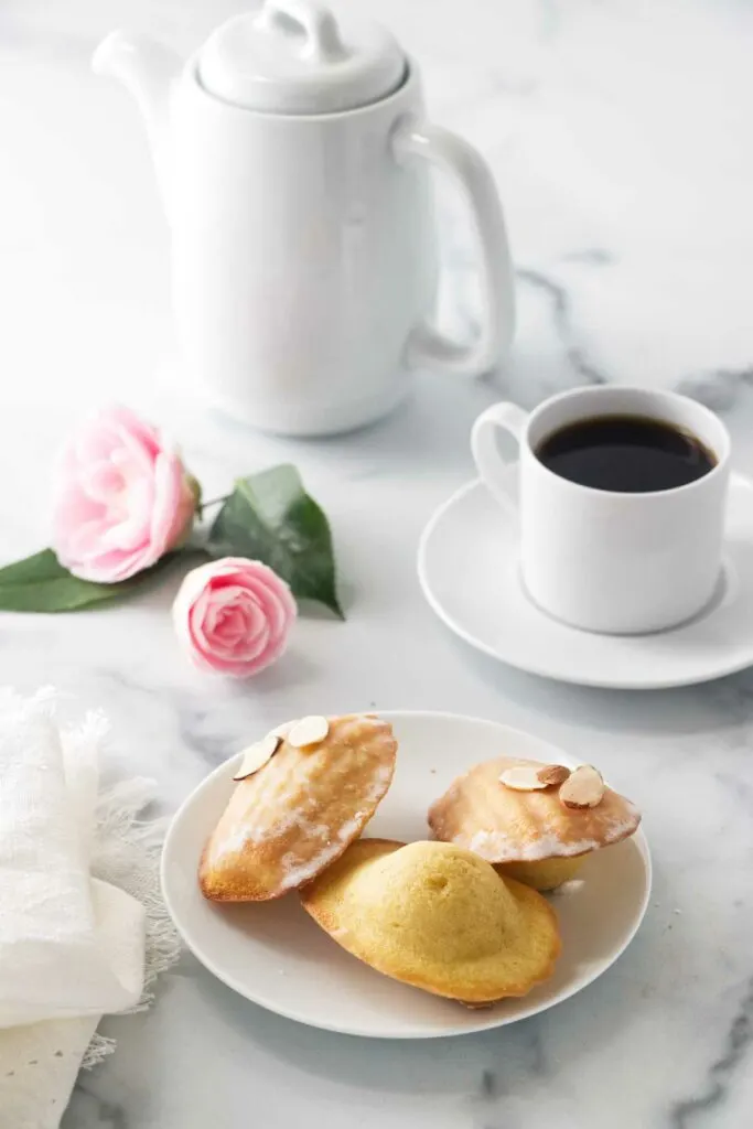 three almond madeleines on a saucer, cup of coffee, coffee pot and two pink camilies.