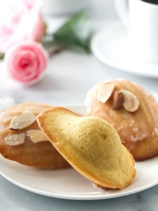 three almond madeleines on a small plate. Coffee cup in the background and a pink rose.