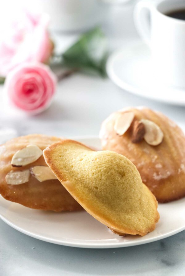 three almond madeleines on a small plate. Coffee cup in the background and a pink rose.