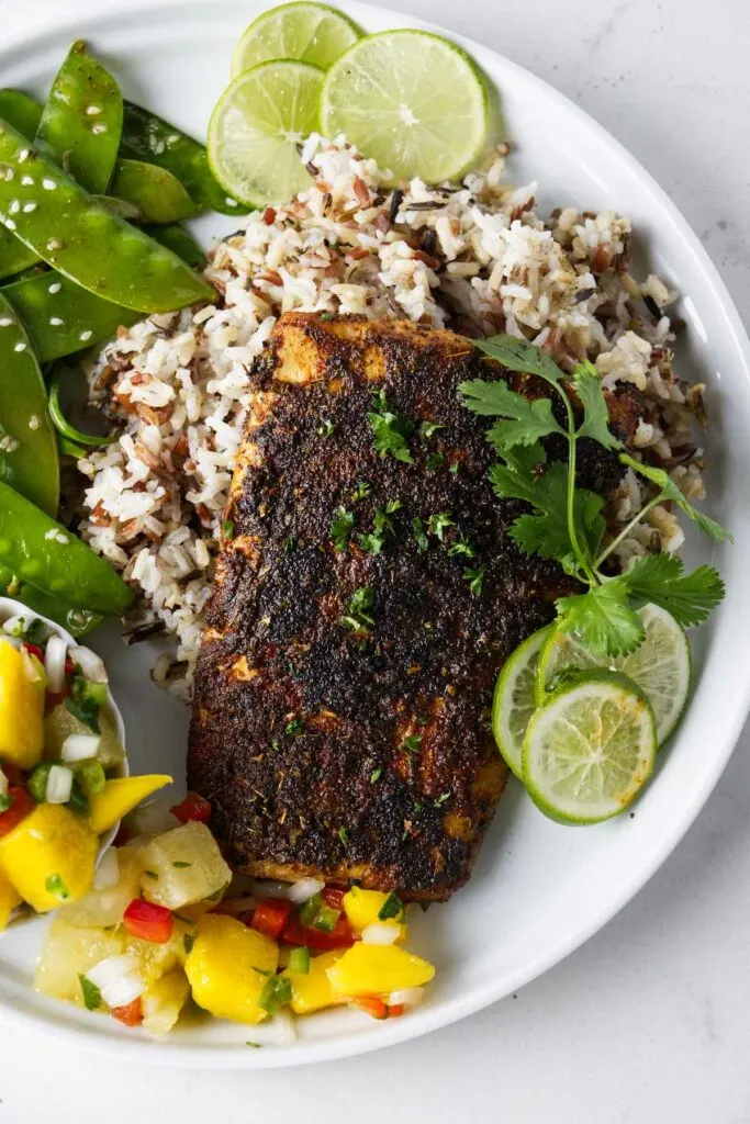 A filet of blackened mahi mahi on a plate with rice, peas, and salsa.