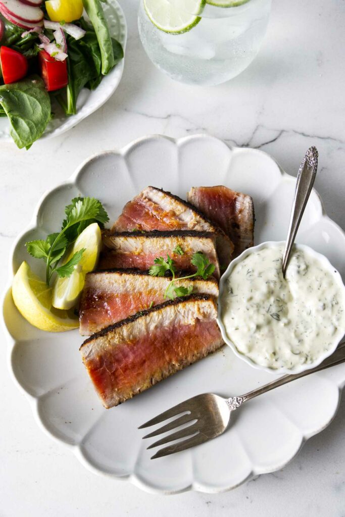 Slices of blackened tuna steak on a plate with aioli sauce on the side next to a green salad.