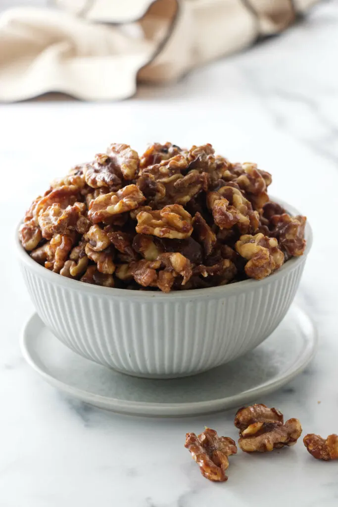 Brown sugar candied walnuts in a dish with several walnuts in the foreground and a towel in the background.