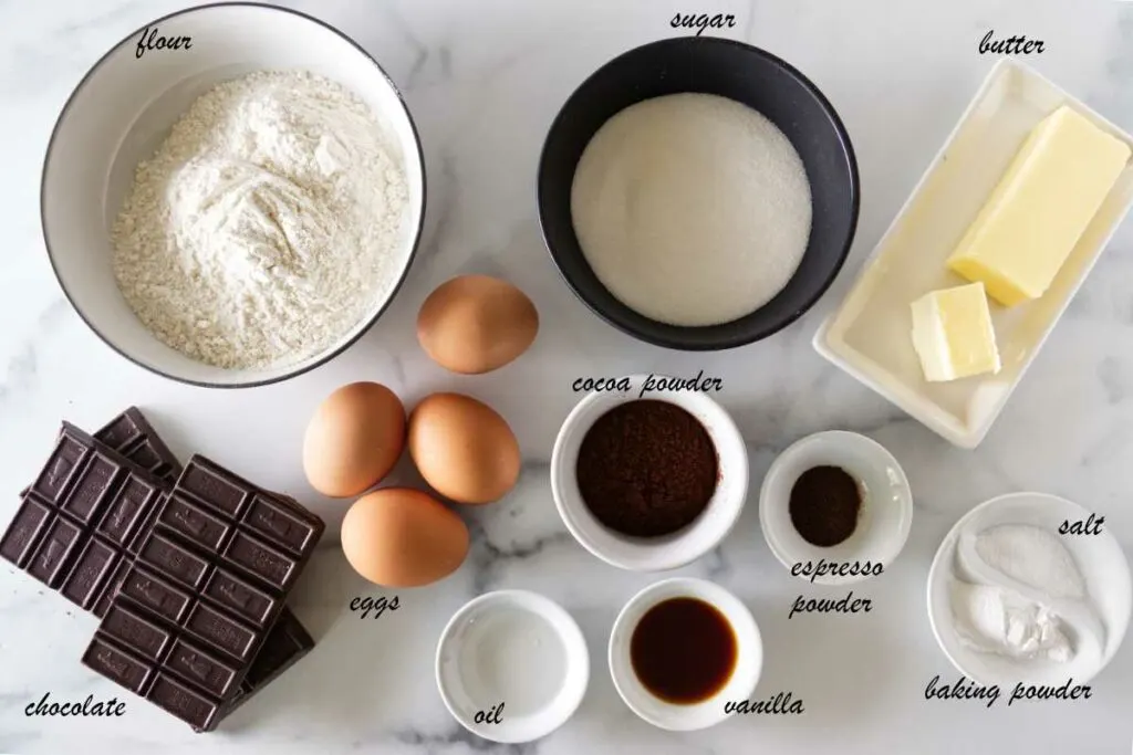 Ingredients needed to make chocolate madeleines, including the chocolate glaze: Flour, sugar, butter, eggs, cocoa powder, espresso powder, baking powder, vanilla, chocolate and oil.