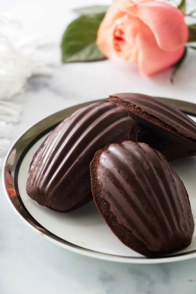 Three chocolate covered madeleines on a plate with a napkin and a flower in the background.