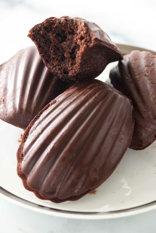 Four chocolate madeleines on a plate with one of the madeleines with a bite out.