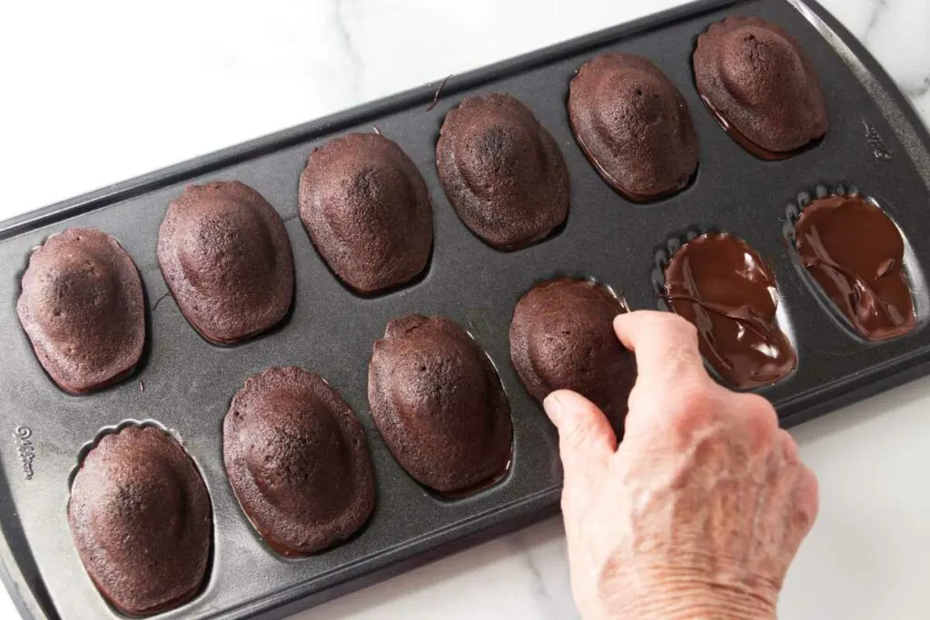 A madeleine pan with baked chocolate madeleines being placed in the chocolate- coated cavities of the pan.