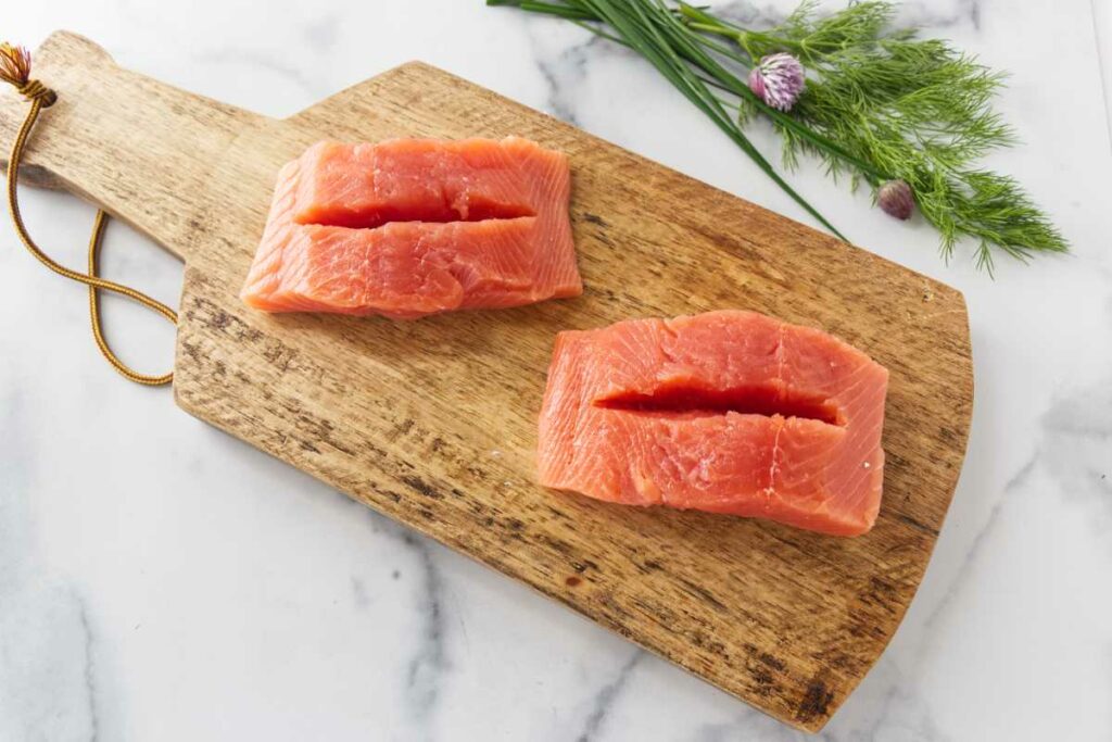 A wood cutting board with two portions of salmon with slits cut for stuffing.