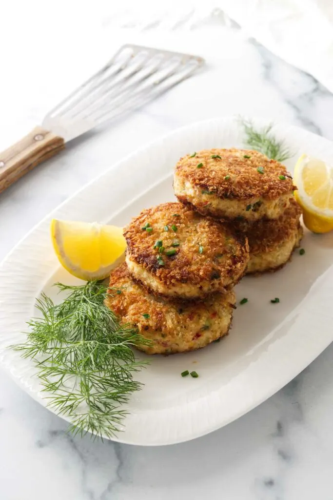 Four servings of crab cakes garnished with snipped chives on a platter, with fresh dill sprigs and lemon wedges. Spatula in the background.