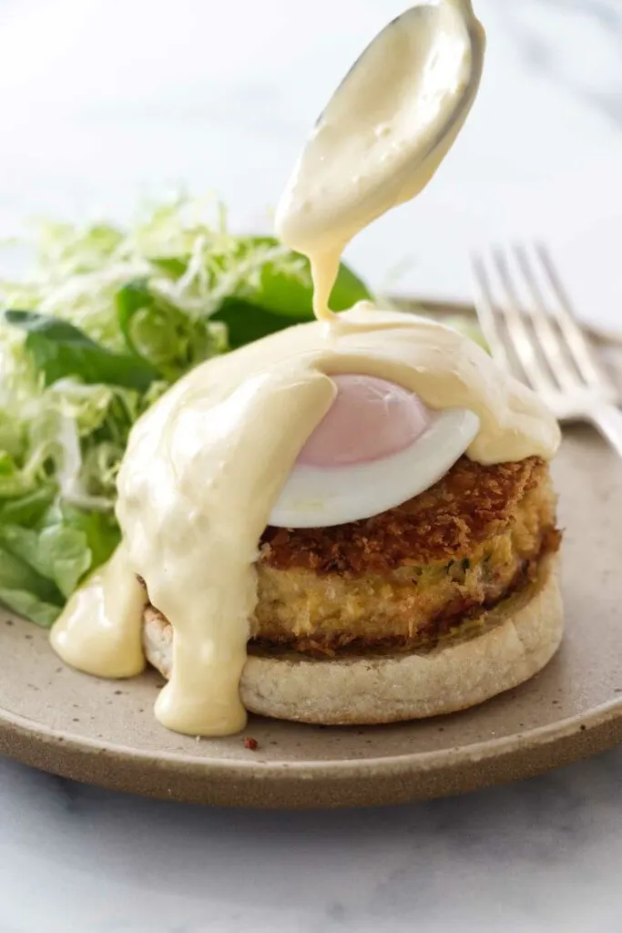 Spooning hollandaise sauce on the poached egg and crab cake eggs benedict. Salad and a fork on the serving plate.