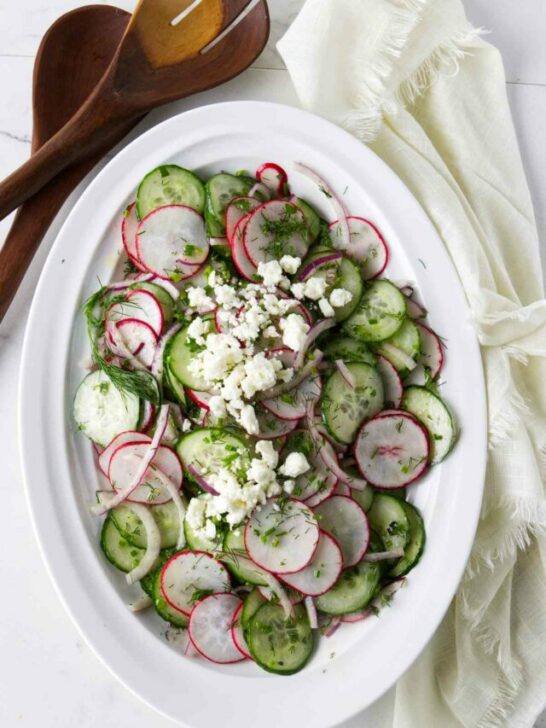 A serving bowl with a cucumber and radish salad next to wooden salad utensils.