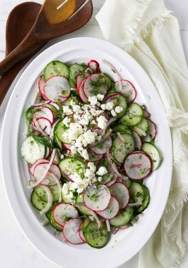 A serving bowl with a cucumber and radish salad next to wooden salad utensils.