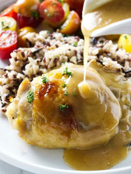 A honey baked chicken thigh on a plate with wild rice and tomatoes. Pouring honey herb sauce over the top.