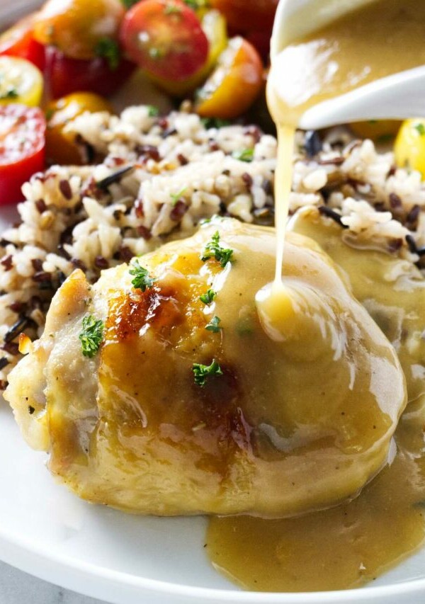 A honey baked chicken thigh on a plate with wild rice and tomatoes. Pouring honey herb sauce over the top.