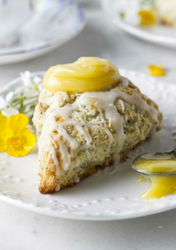 A poppyseed lemon scone on a plate with a spoonful of lemon curd and a tea cup in the background.