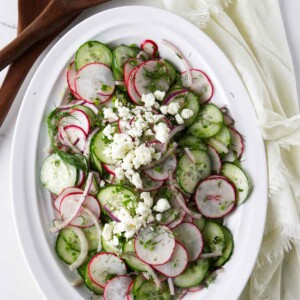 A serving bowl with a cucumber and radish salad next to wooden salad utensils.