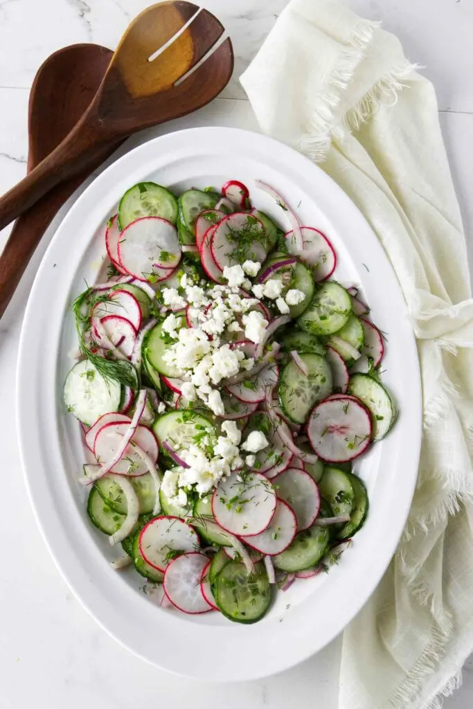 A serving bowl with a cucumber and radish salad next to wooden salad utensils.