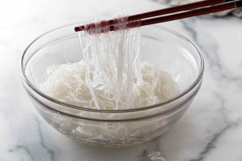 A bowl of cooked, rinsed and drained glass noodles with chop sticks lifting up some noodles.