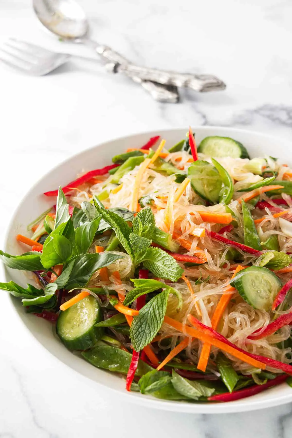 A bowl with tossed glass noodle salad. Salad mixing utensils in the background.
