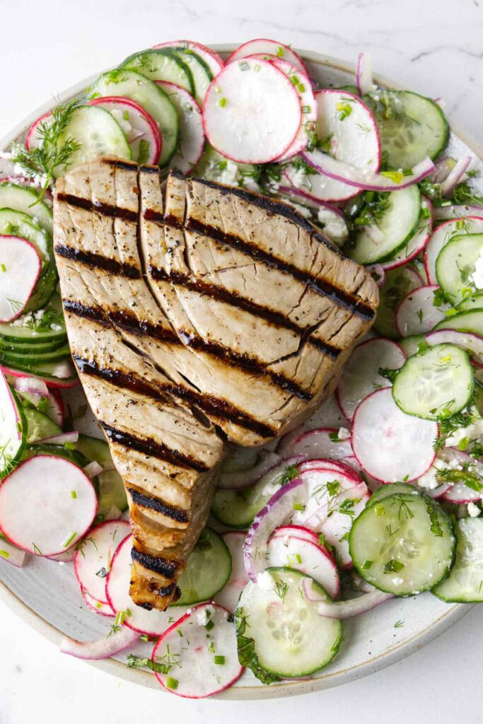 A tuna steak on a plate with a cucumber radish salad.