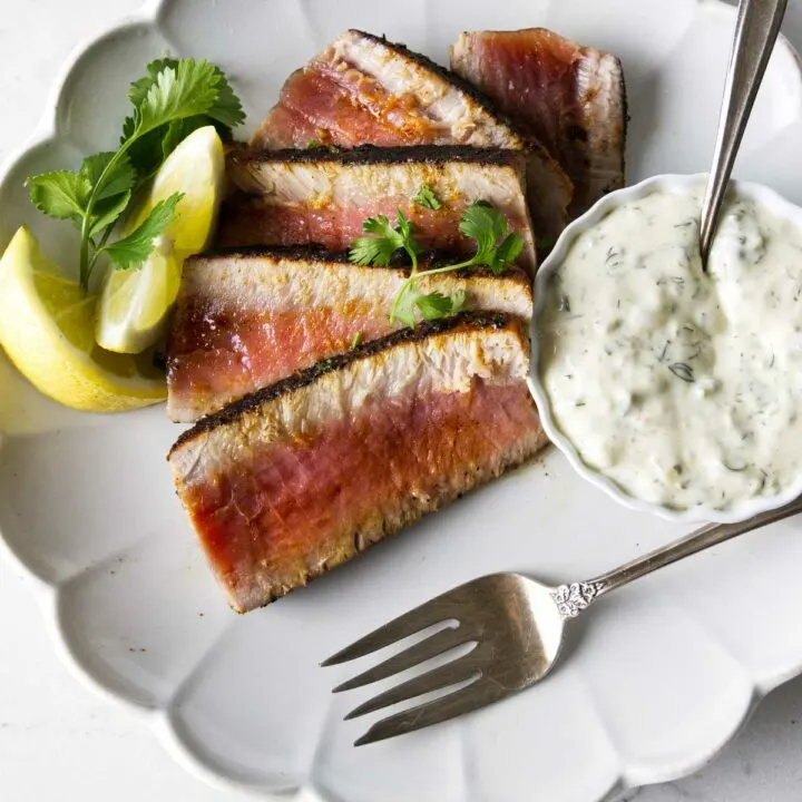 Slices of blackened tuna steak on a plate with lemon slices and aioli sauce on the side.