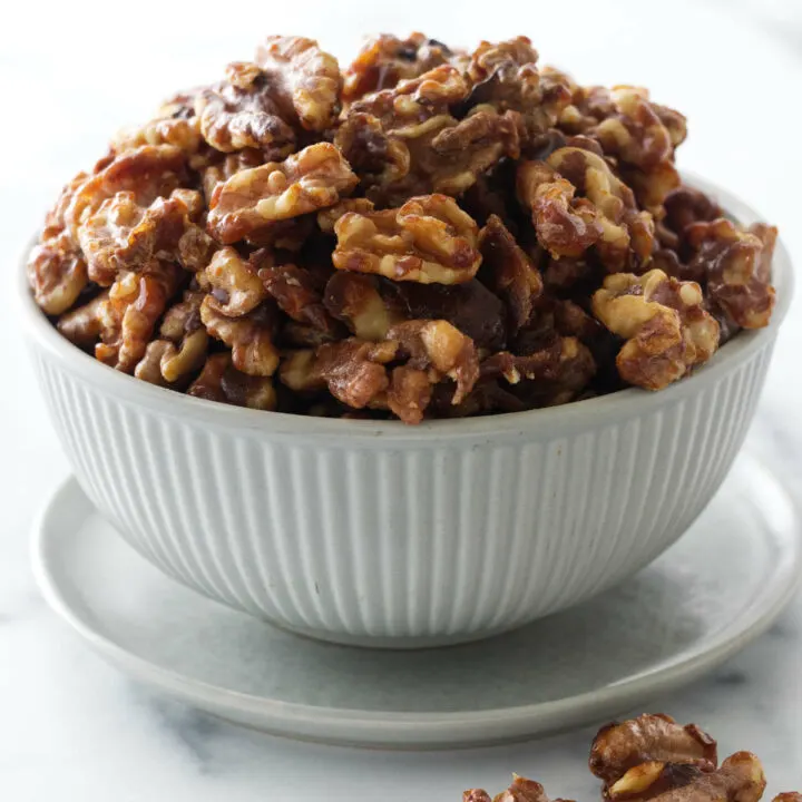 A bowl of candied walnuts on a saucer with several walnuts in the foreground.