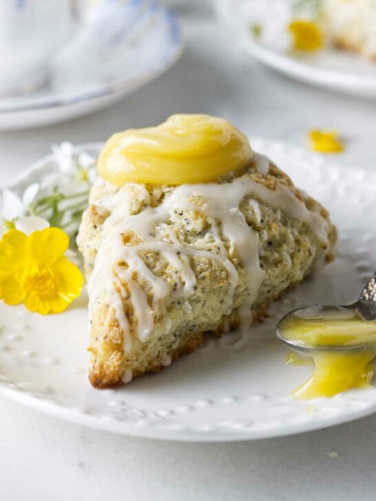 A poppyseed lemon scone on a plate with a spoonful of lemon curd and a tea cup in the background.